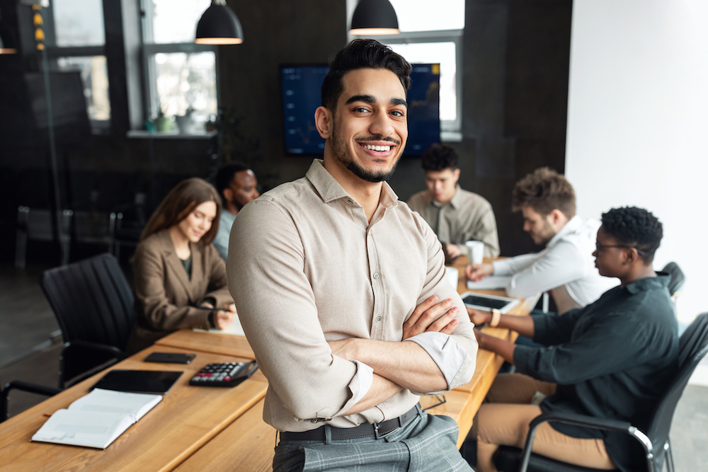 Successful Person. Portrait of confident smiling bearded businessman sitting leaning on desk in office, posing with folded arms and looking at camera, colleagues working in blurred background